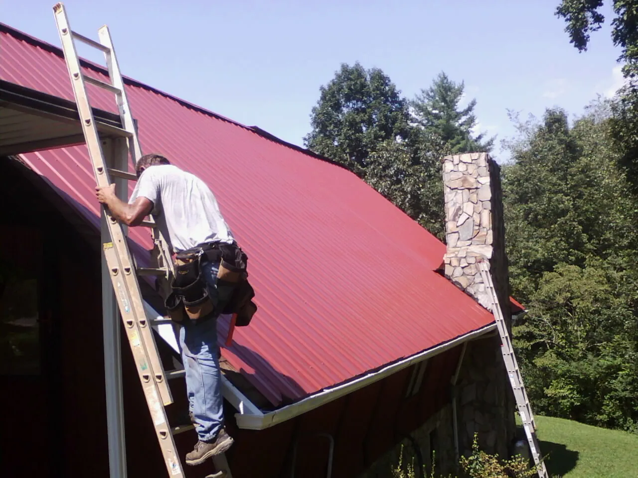 Roofer on a ladder beside a red metal roof and stone chimney — Soffit & Fascia Repair by Roofing Brothers in Iselin